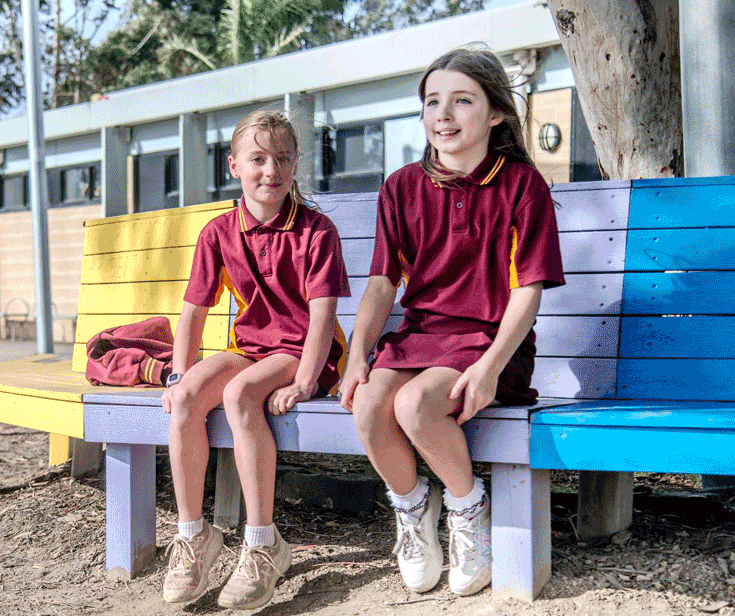 two girls sitting outside in school uniforms on a coloured bench under a gum tree