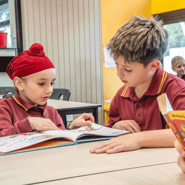 two boys sitting at a table inside a classroom reading a book together