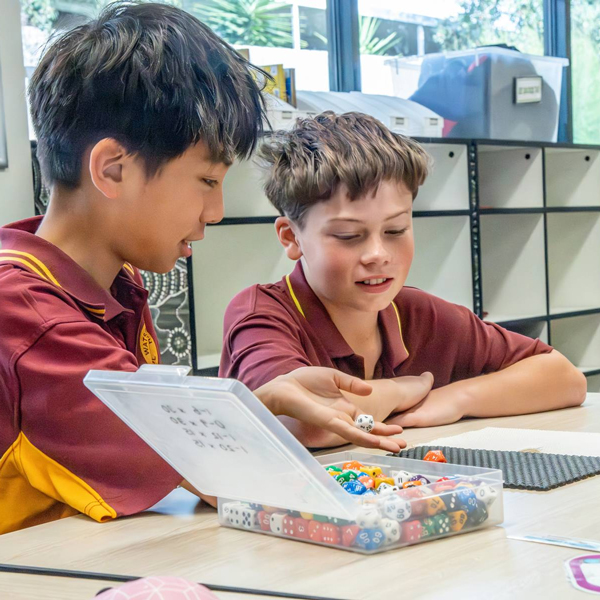 In a classroom, two boys are playing with dice, focused on their game amidst desks and learning resources.