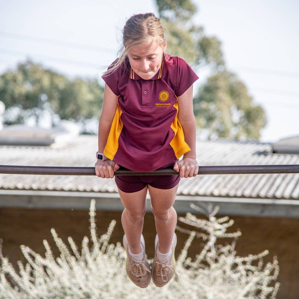 A young girl in a maroon and yellow uniform balances on a bar outdoors. She looks focused, with trees and a corrugated roof in the background.