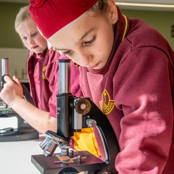 A young girl in a maroon and yellow uniform balances on a bar outdoors. She looks focused, with trees and a corrugated roof in the background.