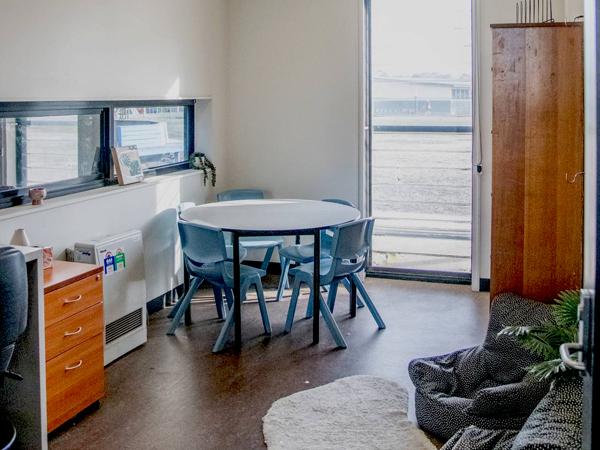 A cozy room with natural light. Features a round table with blue chairs, a wooden cabinet, a window with a view, and a soft rug on a wooden floor.